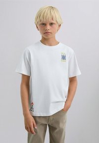 Blonde jongen met hand in broekzak, gekleed in een wit T-shirt met graphic en beige broek, staand tegen een effen lichte achtergrond.