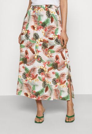 Donna che indossa una gonna bianca con una fantasia floreale tropicale colorata, spacco laterale e sandali verdi, in piedi con le mani nelle tasche.
