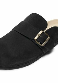 Svart mockaklack med rund tå, med ett enskilt rem och metalldetalj över ovandelen. Mjuk inredning och texturerad sula.