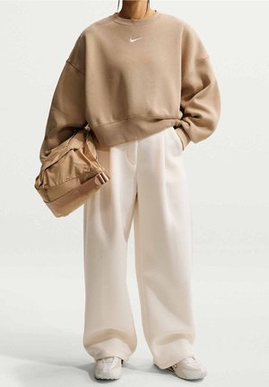 Person iført beige oversized Nike sweatshirt og brede hvide bukser, holder en matchende beige taske i den ene hånd, står stille.