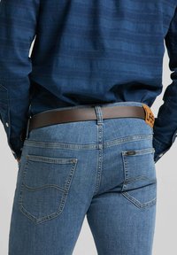 Jean en denim bleu avec une coupe ajustée, présentant des poches arrière et une ceinture en cuir brun. Couture et détails de texture visibles.
