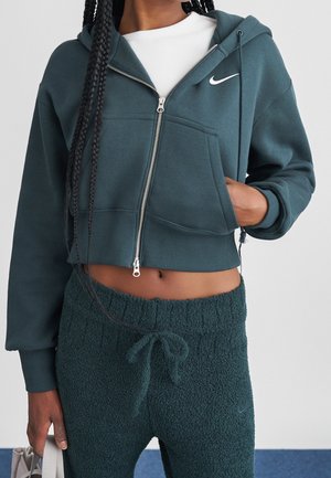 Kvinde iført mørkegrøn cropped lynlås hoodie og matchende teksturerede bukser med snøre, der holder i en bagesnor, stående mod en neutral baggrund.