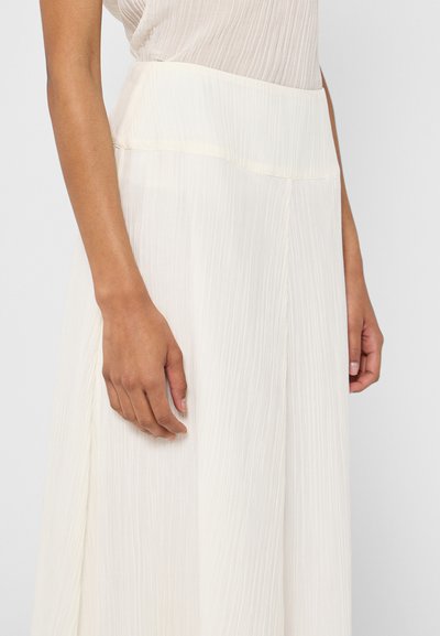 Jupe longue plissée en tissu crème doux et léger. Elle présente une large taille et une silhouette fluide avec des lignes verticales pour la texture.