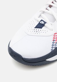 Hvide atletiske sko med mesh-overdel, der har marineblå accenter, røde og blå zigzag mønster, samt gummisål med Nike-logo.