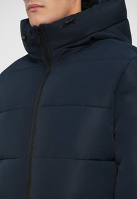 Kario mėlynas pūkinis švarkas su aukštu apykakle, horizontaliais siuviniais, priekiniais užtrauktukais ir reguliuojamais elementais ant gobtuvo.