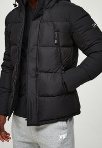 Černá prošívaná bunda s předním zipem, odnímatelnou kapucí a dvěma bočními kapsami. Má prošívanou texturu a jemný detail loga na rukávu.