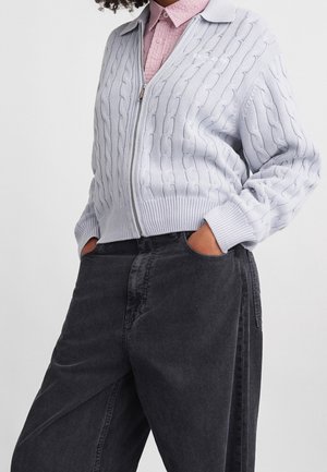 Persona que lleva un suéter gris claro de punto con cremallera y canalé sobre una camisa rosa abotonada, con las manos en los bolsillos de pantalones negros de talle alto y pierna ancha.