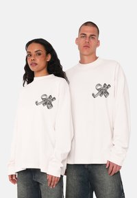 White long-sleeve shirts feature a black graphic design on the chest. The shirts have a relaxed fit and are paired with dark jeans.