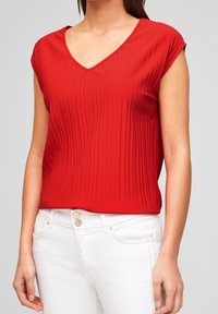 Kvinde iført en rød ribbet kortærmet V-hals top, tisset ind i høj talje hvide jeans med dobbelt dekorative knapper.