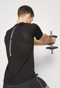 Svart idrottströja med en logotyp på ryggen, tillverkad av lätt material. Tröjan har korta ärmar och en figurnära design.