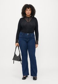 Top preto de manga longa com detalhes em renda no decote e punhos, combinado com jeans azuis de perna larga. Modelo porta uma pequena bolsa preta.