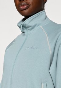 Lyseblå lynlåsjakke med høj krave, med hvid piping og broderet "ONLY PLAY" logo på brystet. Tekstureret stof.