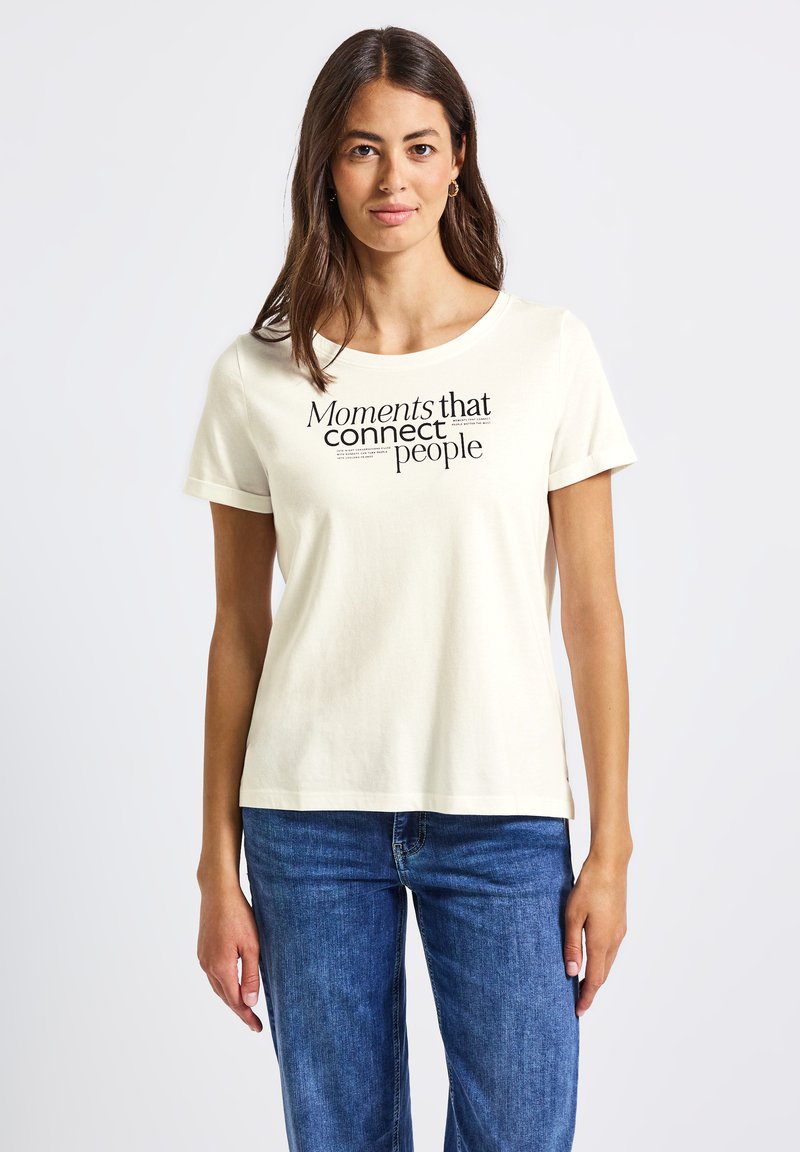 Cremfarbener Baumwoll-T-Shirt mit rundem Halsauschnitt und kurzen Ärmeln. Schwarzer Textdruck "Momente, die Menschen verbinden" in verschiedenen Schriftarten auf der Vorderseite.