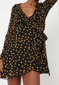 Černé šaty s volánkovým výstřihem a manžetami vyrobené z lehkého materiálu, s hladkým zlatým květinovým vzorem a páskem na zavázání v pase.