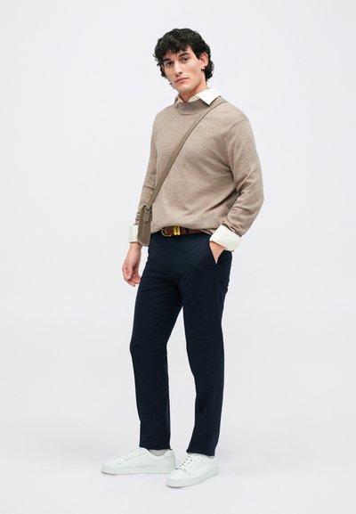 Bežs adīts džemperis ar augstu apkakli, zem tā balta krekla apakškrekls, tumši zili bikses un baltas kedas; nes nelielu plecu somu.