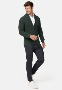Cárdigan de punto acanalado verde oscuro con cierre de cremallera, llevado sobre una camisa blanca con cuello, combinado con pantalones oscuros y zapatillas blancas.