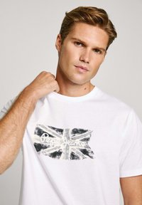 Biała bawełniana koszulka z krótkim rękawem, z wyblakłym nadrukiem flagi Union Jack oraz tekstem "Pepe Jeans London" w wyraźnej czerni i złocie.