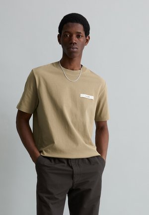 Jonge man met kort krullend haar, gekleed in een beige T-shirt met een klein embleem en zwarte broek, staand met handen in de zakken.
