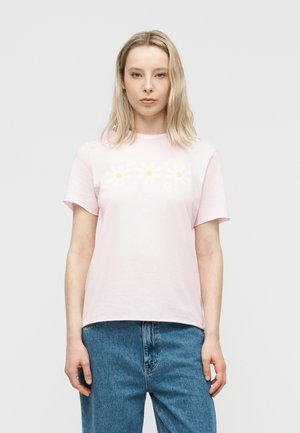 Młoda kobieta o prostych, blond włosach, ubrana w jasnoróżową koszulkę z białymi stokrotkami i niebieskie dżinsy, stojąca na tle jednolitego tła.