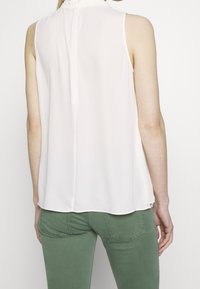 Bezrękawnik, biały blouse wykonany z lekkiego materiału, z koronkowym kołnierzykiem i detalem szwu z tyłu. W zestawie z zielonymi spodniami.