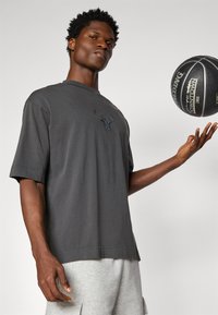 Grå bomulls t-skjorte med korte ermer, med et svart okse-trykk på brystet. Modellen holder en basketball i den ene hånden.