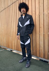 Svart sportoverall med vita accenter; lättviktsmaterial, zip-up jacka, smalare byxor och sportskor. Träbakgrund.