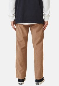 Person som bär beige kordflöjlsbyxor, svarta sneakers med vita sulor och lager-på-lager långärmade toppar i svart och vitt, står vänd bort.