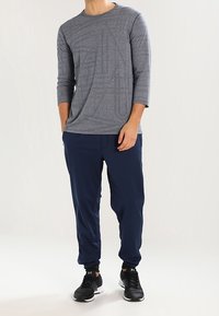 Man som bär en grå långärmad skjorta, marinblå joggingsbyxor och svarta sneakers, stående med ena handen i fickan.