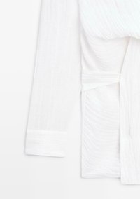 Weißes, strukturiertes Wickelkleid mit langen Ärmeln, breiten Manschetten und einem Stoffgürtel, der an der Taille gebunden ist.