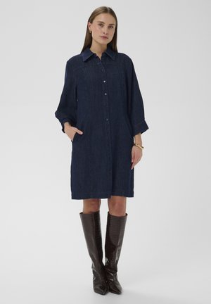 Donkerblauwe denim overhemdjurk met kraag, knoopsluiting aan de voorkant en zakken. Heeft lange mouwen en valt boven de knie. Bruine leren laarzen tot aan de knie.