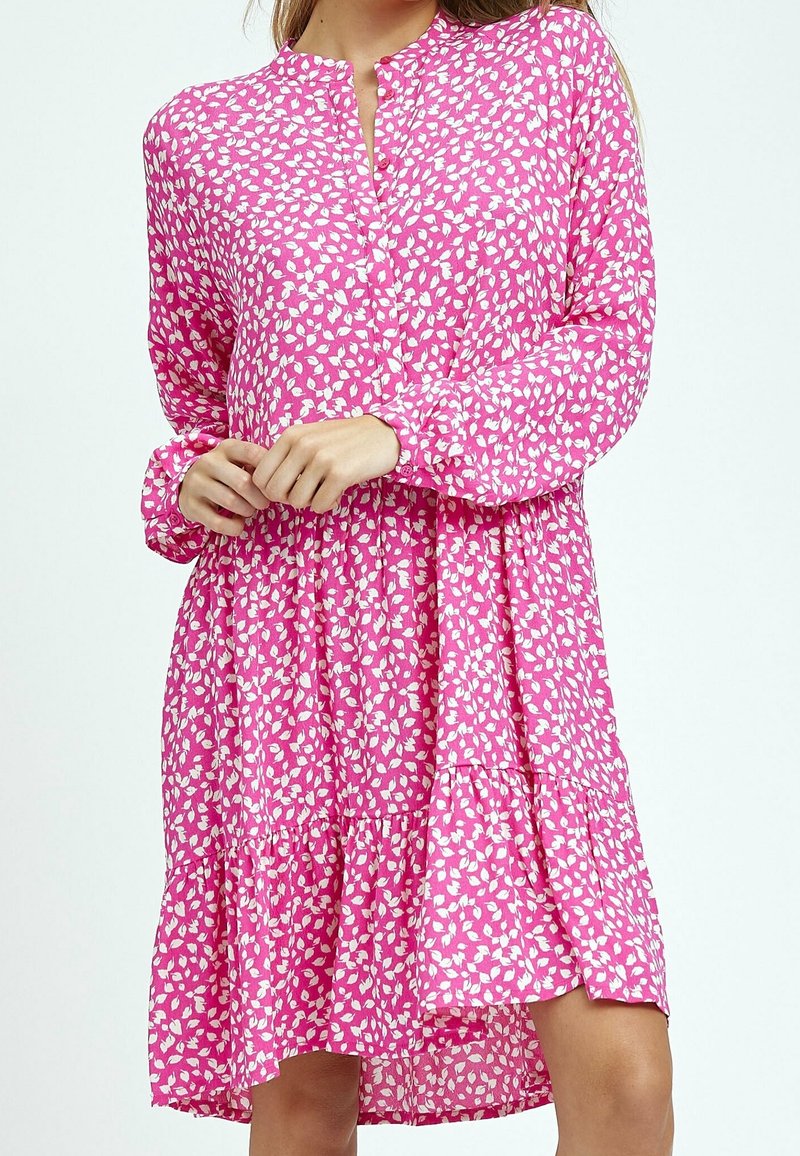 Femme portant une robe rose à manches longues avec un motif de feuilles blanches, jupe froncée et petite fente en V, vue du cou jusqu'à la mi-cuisse.