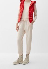 Gilet matelassé rouge, haut à manches longues beige clair, jogging beige avec poignets élastiques, et bottes beiges à lacets avec surface texturée et accents métalliques.