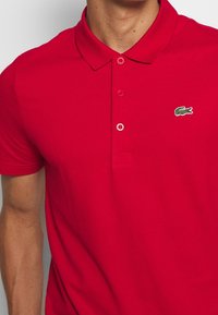 Red polo shirt made of cotton, featuring a classic collar, three buttons, and an embroidered green crocodile logo on the chest.