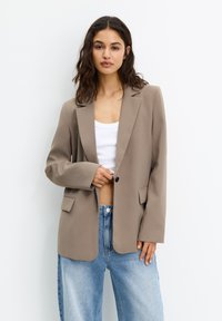 Blazer beige en tissu lisse, avec un revers cranté, fermeture à un bouton et deux poches avant. Porté par-dessus un haut blanc avec un jean bleu.