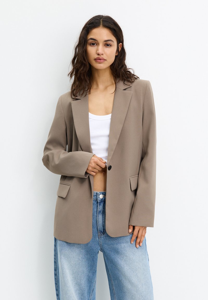 Blazer beige en tissu lisse, avec un revers cranté, fermeture à un bouton et deux poches avant. Porté par-dessus un haut blanc avec un jean bleu.