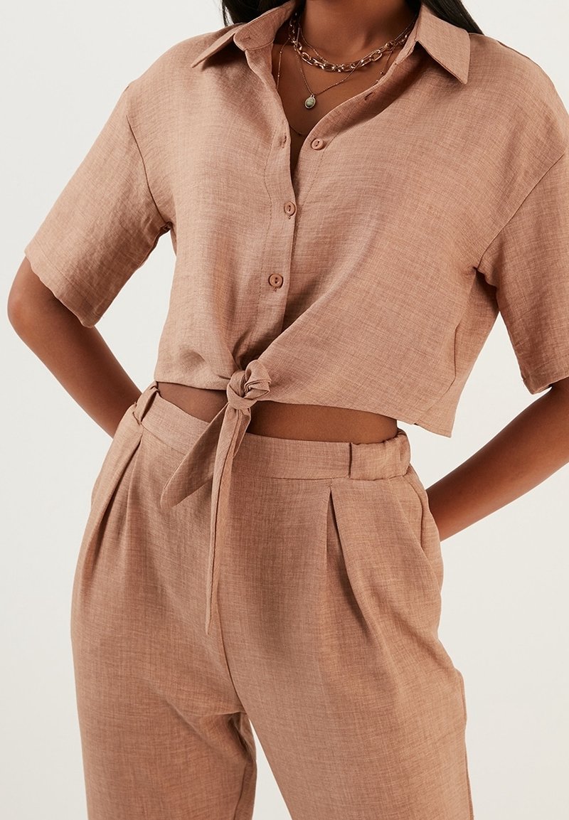Femme portant une chemise marron clair à manches courtes boutonnée et nouée à la taille, et un pantalon plissé taille haute assorti avec des poches latérales.
