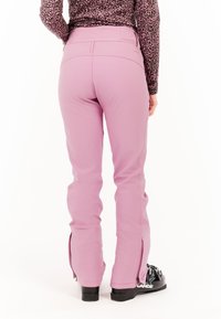 Ružové snehové nohavice so zúženým strihom, vyrobené z hladkej látky. Obsahujú bočné zipsy na členkoch a sú nosené s čiernymi lyžiarskymi topánkami.