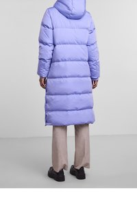 Lång puffjacka i ljuslila med huva, med horisontella quiltade sektioner och sidoklyftor för enkel rörelse. Stylerad med beige byxor.