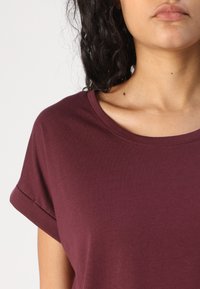 Tricou burgund cu guler rotund, mâneci scurte rulate și material din bumbac moale, prezentând un design simplu cu textură netedă.