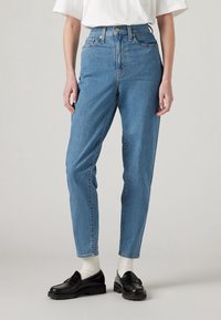 Personne portant un jean bleu taille haute, des chaussettes blanches, des mocassins noirs et une chemise blanche à manches courtes, debout devant un fond uni.