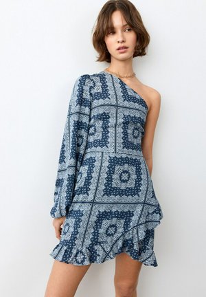 Giovane donna che indossa un vestito monospalla blu e bianco con manica lunga e orlo arricciato, in piedi davanti a uno sfondo bianco.