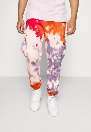 Mężczyzna noszący wielokolorowe spodnie typu cargo w stylu tie-dye w kolorach czerwonym, pomarańczowym, fioletowym i białym, w białych adidasach na szarej podłodze.