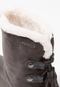 Botte en suede gris foncé avec doublure en fausse fourrure blanche, lacets décoratifs et accents métalliques. Présente le logo "TOM TAILOR" sur le côté.