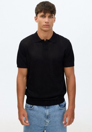 Joven con cabello corto castaño que lleva una camisa polo de punto negra de manga corta y vaqueros azul claro, de pie frente a un fondo liso.