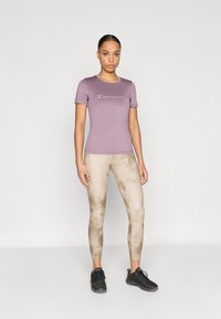 Μωβ αθλητική κοντομάνικη μπλούζα με το λογότυπο "Champion", σε συνδυασμό με μπεζ κολάν με σχέδιο ταϊ-ντάι και μαύρα αθλητικά παπούτσια.