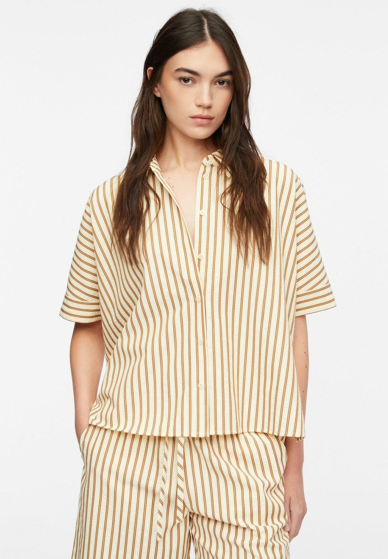Jeune femme aux longs cheveux bruns portant une chemise beige ample et un pantalon à rayures verticales marron, debout devant un fond blanc uni.