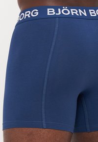 Navy blue boxer shorts made of soft fabric with a fitted shape, featuring a white branded waistband and contrasting blue stitching details.