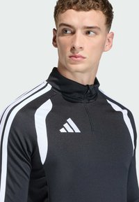 Μαύρη αθλητική μπλούζα με μισό φερμουάρ, άσπρες λωρίδες στα πλάγια και λογότυπο της μάρκας. Λείο υλικό, εφαρμοστή σχεδίαση, υψηλή λαιμόκοψη και αναπνεύσιμο ύφασμα.