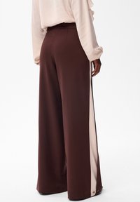 Osoba ubrana w brązowe spodnie wide-leg z beżowymi paskami po bokach i zatrzaskami, w połączeniu z jasnobeżową bluzką z długim rękawem, stojąca bokiem.