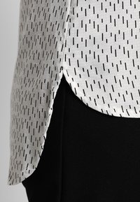 Blouse blanche avec motif rayé vertical noir, présentant un ourlet arrondi et des fentes latérales. Portée sur un matériau noir, mettant en avant le contraste des couleurs.